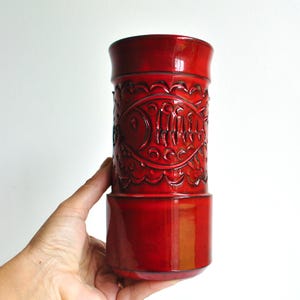 May include: A glossy red ceramic vase with a cylindrical body and a flared top. The vase features an embossed design of fish and other decorative elements. The vase is held against a white background.