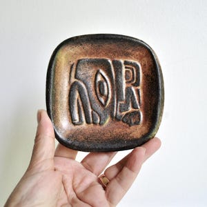 May include: A square, brown ceramic dish with a raised, abstract design. The dish has a textured surface and a dark brown border with lighter brown and yellow accents. The design appears to be a stylized face or figure.