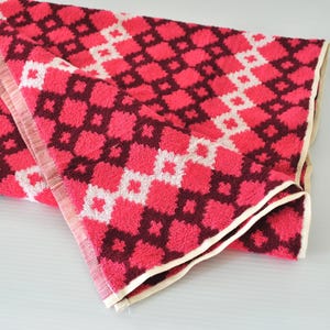 May include: A folded rectangular towel featuring a repeating geometric pattern of dark maroon and bright pink squares, accented by a diagonal stripe of white diamond shapes. The edges of the towel are finished with a simple, light beige fabric trim.