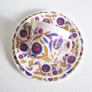 May include: A white teacup and saucer set with a floral pattern in shades of purple, blue, and gold. The pattern features stylised flowers and leaves.