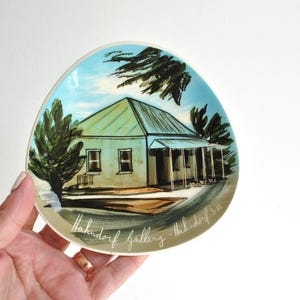 May include: A small, oval decorative plate with a hand-painted image of a house with a green roof and porch. The plate has a blue sky background, green trees, and the text "Hahndorf gallery - Hahndorf SA".