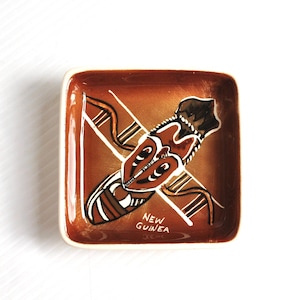 May include: A square ceramic dish with a glossy brown glaze featuring a hand-painted design of a mask and abstract lines in white, black, and tan. The text NEW GUINEA is printed in white at the bottom centre of the dish, which has rounded corners.