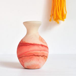 May include: A small, round vase with a narrow neck and a flared rim. The surface features a marbled pattern with swirling shades of coral, peach, and cream, creating a smooth, matte appearance across the spherical body of the vessel.