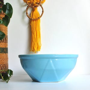 May include: A bright light blue ceramic bowl with a faceted geometric exterior design. The bowl features a smooth, glossy finish and a wide, circular rim, showcasing a modern, angular shape that tapers down to a smaller, flat hexagonal base.