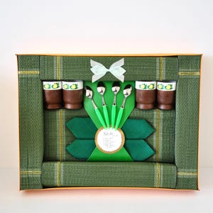 May include: A gift set featuring four brown cups with yellow floral rims, four silver spoons with bright green handles, and four dark green napkins. The items are arranged in a green woven box with a white ribbon bow and a circular label.