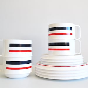 May include: Set of white ceramic mugs with black and red stripes. The mugs are stacked on top of each other. There are also white ceramic plates stacked on top of each other.
