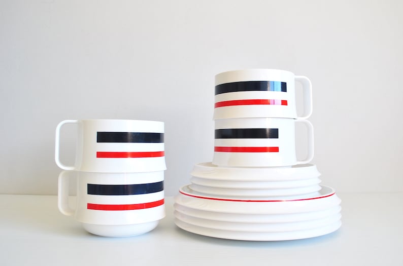 May include: Set of white ceramic mugs with black and red stripes. The mugs are stacked on top of each other. There are also white ceramic plates stacked on top of each other.