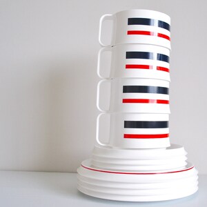 May include: A stack of four white ceramic mugs with black and red stripes. The mugs are stacked on top of a set of white ceramic plates.