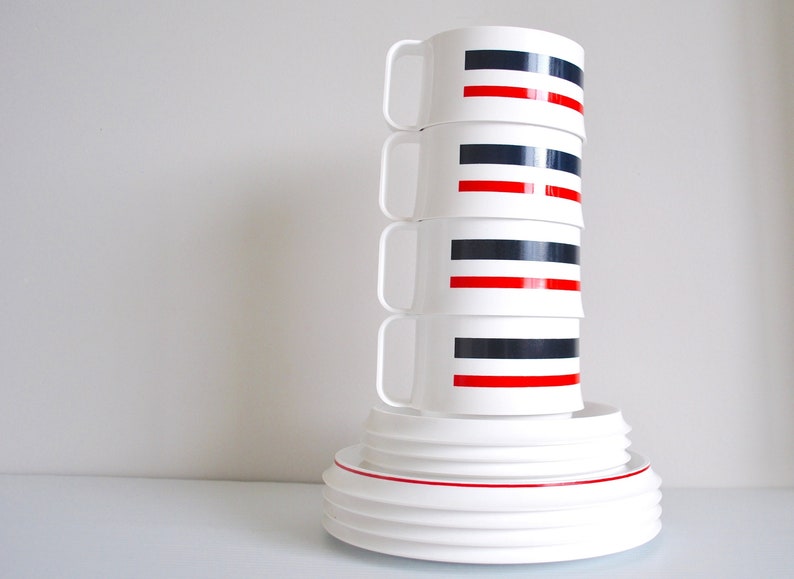 May include: A stack of four white ceramic mugs with black and red stripes. The mugs are stacked on top of a set of white ceramic plates.