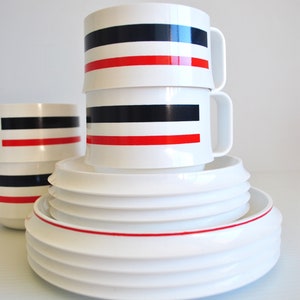 May include: A set of white ceramic mugs with red and black stripes stacked on top of each other. The mugs are on top of a set of white ceramic plates with a red rim.