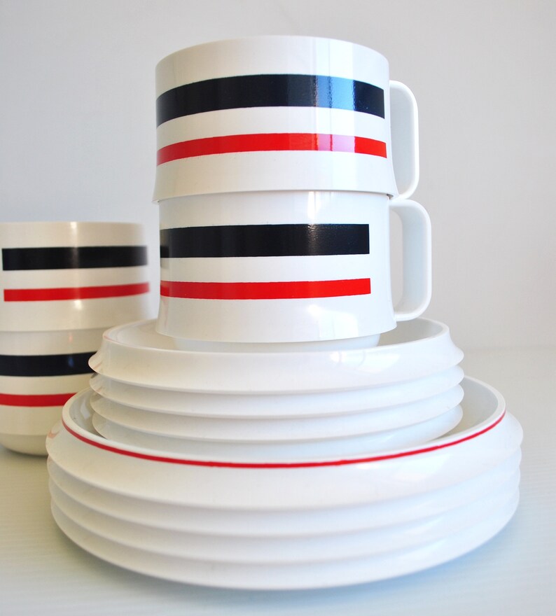 May include: A set of white ceramic mugs with red and black stripes stacked on top of each other. The mugs are on top of a set of white ceramic plates with a red rim.