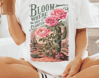 May include: White t-shirt with a graphic print of pink roses growing from a cactus in a desert landscape. The text "Bloom Where You Are Planted" is printed in green on the shirt.