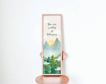 May include: A framed print featuring a watercolour illustration of mountains and trees. The text "You are worthy of belonging" is written in green cursive on a pale beige background.