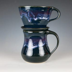 May include: A two-piece ceramic coffee set with a dark blue and purple glaze. The top piece is a pour-over coffee maker, and the bottom is a mug with a handle. The set is designed for brewing and enjoying coffee.