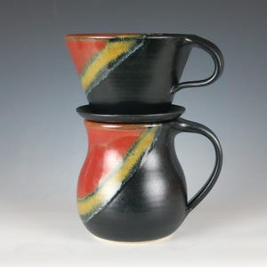 May include: A handmade ceramic coffee pour-over set. The set includes a black mug with a handle, a matching pour-over cone, and a small black saucer. The mug and cone have a diagonal design in red, yellow, and white.