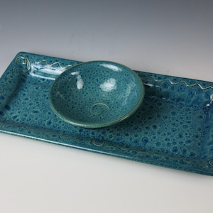 May include: A teal ceramic serving set featuring a rectangular tray and a small round bowl. The set has a textured surface and gold-colored decorative accents along the edges. The bowl sits on the tray.