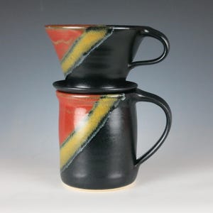 May include: A ceramic pour-over coffee maker set. The set includes a black mug with a handle, a matching pour-over cone, and a small plate. The mug and cone have a diagonal design in red and yellow.