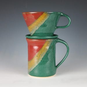 May include: Handmade ceramic coffee pour-over set. The set includes a green mug with a handle, a matching pour-over cone, and a base. The mug and cone feature a diagonal design in red, yellow, and green. The set is approximately 6 inches tall.