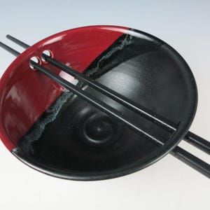 May include: A ceramic ramen bowl with a two-tone design, featuring a red and black color scheme. The bowl has a circular shape with two holes for chopsticks. Black chopsticks are placed across the bowl.