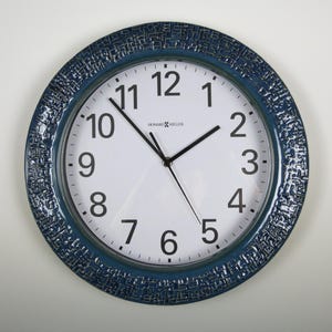 May include: Round wall clock with a white face, black numerals, and black hands. The clock has a textured, dark blue bezel. The brand name "HOWARD MILLER" is printed on the face.