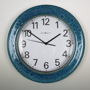 May include: Round wall clock with a white face and black numerals. The clock has black hands and a textured, teal blue bezel. The brand name "HOWARD MILLER" is printed on the face.