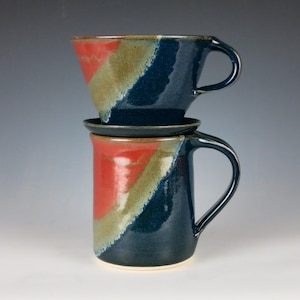 May include: Handmade ceramic coffee pour-over set. The set includes a mug, a pour-over cone, and a small plate. The mug and cone are navy blue with red and tan diagonal stripes. The set is designed for brewing coffee.