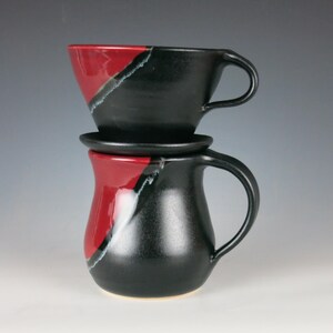 May include: A black and red ceramic pour-over coffee set. The set includes a carafe with a handle, a filter holder, and a small saucer. The design features a diagonal color block of red and black with a white accent.