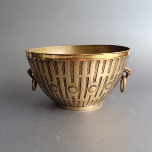 May include: A gold-colored metal bowl with a decorative pattern of vertical lines and circles. The bowl has two small, round handles on the sides.