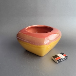 May include: A ceramic bowl with a square base and rounded top, featuring a gradient of colors: yellow, brown, and pink. A small matchbox is next to the bowl. The bowl is approximately 4 inches wide.