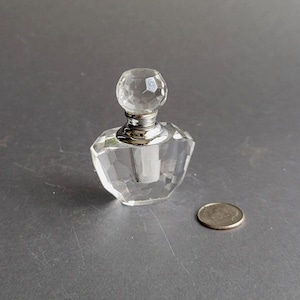 May include: A clear glass perfume bottle with a faceted design and a silver cap. The bottle has a faceted stopper. The bottle is sitting on a gray surface.