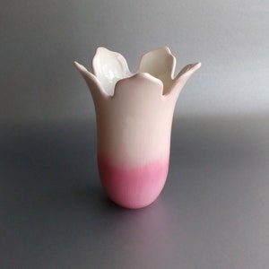 May include: A white ceramic vase with a pink ombre glaze. The vase has a tulip-shaped body with a wide, scalloped opening.