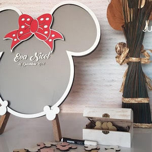 May include: White wooden sign shaped like Mickey Mouse ears with a red bow and white polka dots. The sign has a gray chalkboard surface with the text "Eva Nicol 21 December 2018".