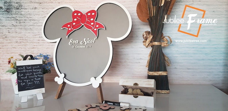 May include: White wooden sign shaped like Mickey Mouse ears with a red bow and white polka dots. The sign has a gray chalkboard surface with the text "Eva Nicol 21 December 2018".