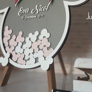 May include: A gray wooden Mickey Mouse shaped sign with a white border and pink and white Mickey Mouse ears inside. The sign has the text "Eva & Nicol" and "26 Decembrie 2018" written on it. The sign is on a wooden easel. A small chalkboard sign with the text "write hear you'r wellcome mesage for your guests and invite them to sign a heart for you!!!" is in the foreground. A white wooden box with a hinged lid is open and contains small wooden Mickey Mouse ears. The box is on a wooden surface. The text "Jubilee Frame" and "www.jubileetree.com" is visible in the background.