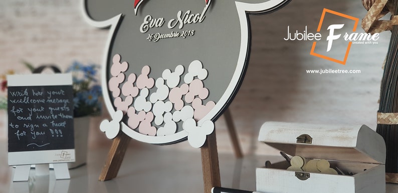May include: A gray wooden Mickey Mouse shaped sign with a white border and pink and white Mickey Mouse ears inside. The sign has the text "Eva & Nicol" and "26 Decembrie 2018" written on it. The sign is on a wooden easel. A small chalkboard sign with the text "write hear you'r wellcome mesage for your guests and invite them to sign a heart for you!!!" is in the foreground. A white wooden box with a hinged lid is open and contains small wooden Mickey Mouse ears. The box is on a wooden surface. The text "Jubilee Frame" and "www.jubileetree.com" is visible in the background.