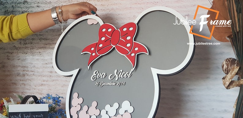 May include: A gray and white wooden frame shaped like Mickey Mouse ears with a red bow and white polka dots. The frame has a gray background with the text "Eva Nicol" and "26 Decembrie 2018" written in white. The frame is filled with small white and pink Mickey Mouse ears.