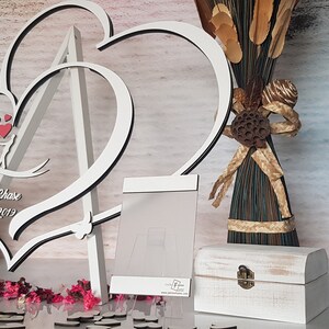 May include: White wooden double heart frame with a stand, personalized with the names "Jonathan & Chase" and the date "7-4-2019". The frame is decorated with small red hearts and is sitting on a table with a white wooden box and a small bouquet of flowers.