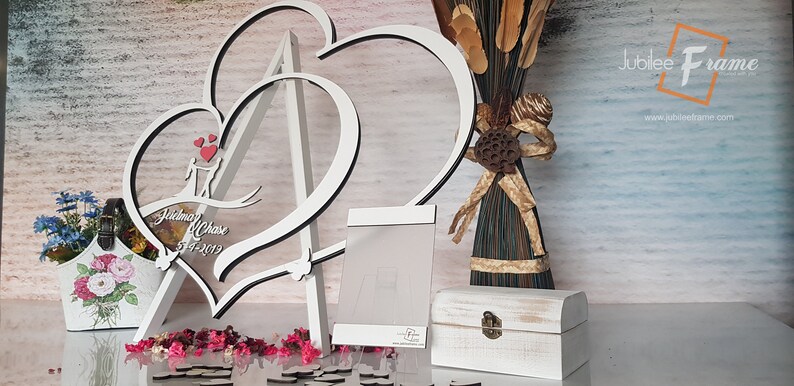 May include: White wooden double heart frame with a stand, personalized with the names "Jonathan & Chase" and the date "7-4-2019". The frame is decorated with small red hearts and is sitting on a table with a white wooden box and a small bouquet of flowers.