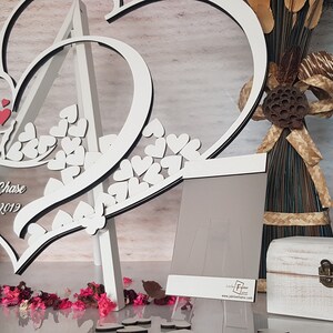 May include: White wooden double heart cutout with a clear acrylic center. The hearts are decorated with small white hearts and the text "Jacqueline & Chase 5-4-2019". The hearts are standing on a white easel.