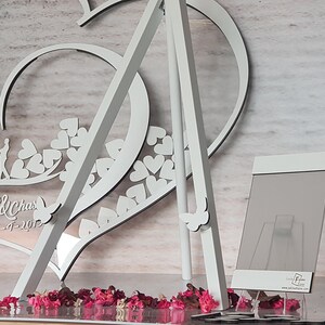 May include: White wooden heart-shaped wedding sign with the names "Jeielma & Chas" and the date "5-4-2017" engraved on it. The sign is standing on a white easel and surrounded by pink and red flower petals.