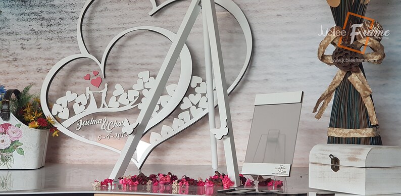 May include: White wooden heart-shaped wedding sign with the names "Jeielma & Chas" and the date "5-4-2017" engraved on it. The sign is standing on a white easel and surrounded by pink and red flower petals.