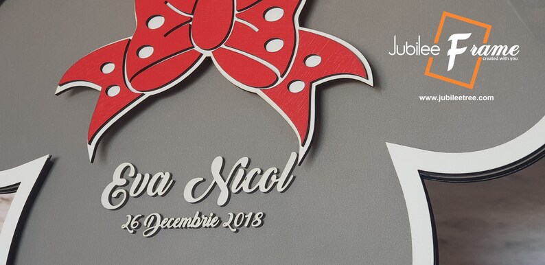 May include: A gray wooden frame with a white border and a red bow with white polka dots. The frame has a cutout in the shape of a cloud with the text "Eva Nicol" and "26 Decembre 2018" written in white. The frame is from Jubilee Frame, created with you.