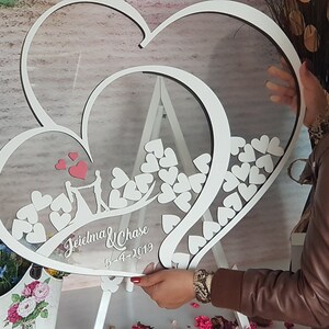 May include: White wooden double heart shaped frame with a clear acrylic insert. The frame is decorated with small white hearts and a couple silhouette. The text "Jeidlma & Chase 5-4-2019" is written on the frame.