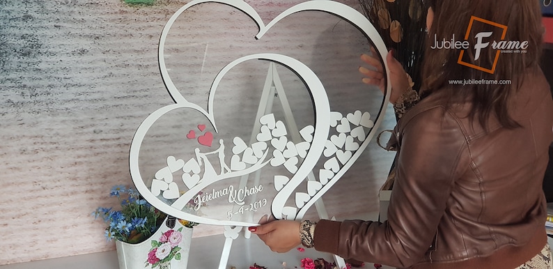 May include: White wooden double heart shaped frame with a clear acrylic insert. The frame is decorated with small white hearts and a couple silhouette. The text "Jeidlma & Chase 5-4-2019" is written on the frame.