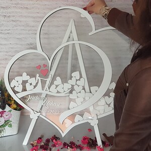 May include: White wooden double heart frame with a clear acrylic insert. The frame is standing on a white easel and has a couple silhouette and the text "Jaidyn & Chase 5-1-2023" inscribed on the bottom. The frame is filled with small white hearts.