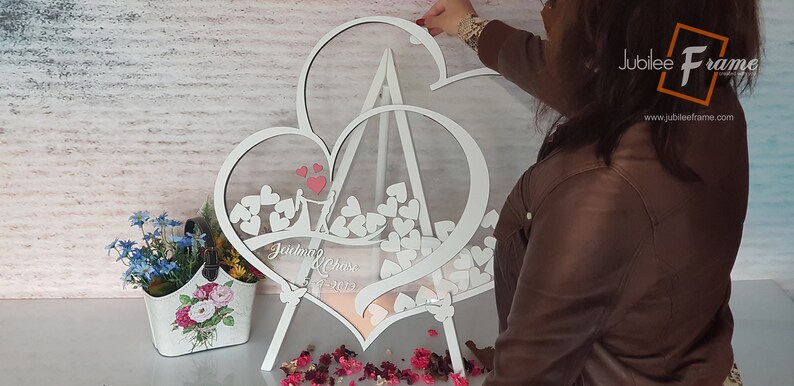 May include: White wooden double heart frame with a clear acrylic insert. The frame is standing on a white easel and has a couple silhouette and the text "Jaidyn & Chase 5-1-2023" inscribed on the bottom. The frame is filled with small white hearts.
