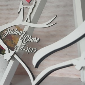 May include: White wooden heart-shaped frame with a black outline. The frame features a cutout of a couple holding hands and a ribbon with the text "Jeielma & Chase 5-4-2019".