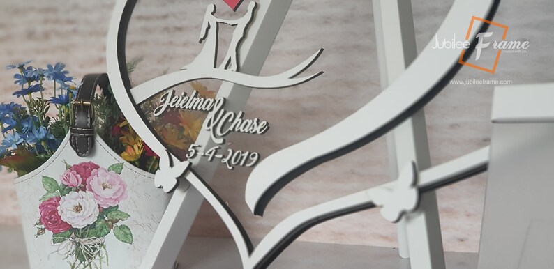 May include: White wooden heart-shaped frame with a black outline. The frame features a cutout of a couple holding hands and a ribbon with the text "Jeielma & Chase 5-4-2019".