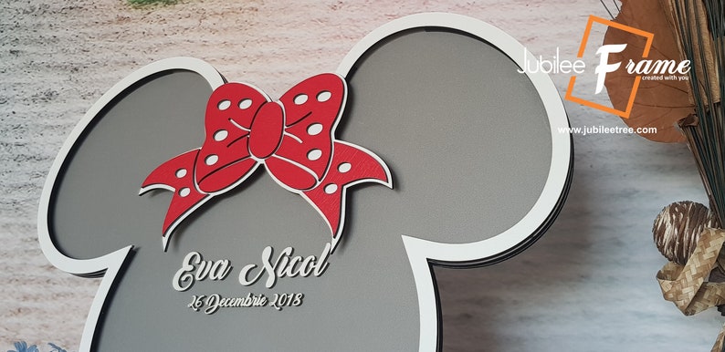 May include: A gray and white wooden cutout of a Mickey Mouse head with a red bow with white polka dots. The cutout has the text "Eva Nicol" and "26 December 2018" written on it.