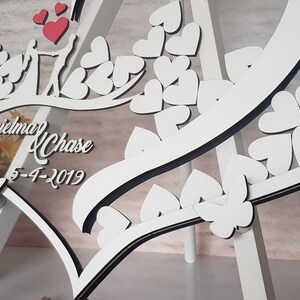 May include: White wooden heart-shaped sign with a clear acrylic insert. The sign features a couple's names, "Jeri and Chase", and their wedding date, "5-4-2019". The sign is decorated with small white hearts and a black border.